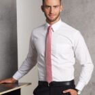 A man wearing a white dress shirt with a pink tie and black pants leans against a countertop. He is standing against a gray background and is smiling at the camera.