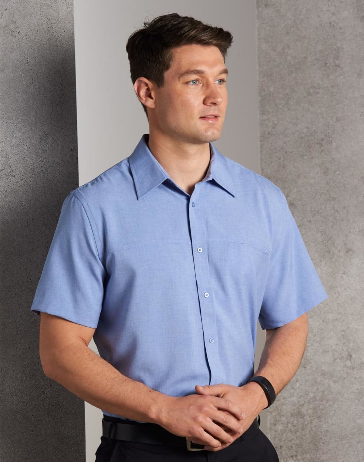 A man wearing a light blue short-sleeved button-up shirt stands against a gray wall, looking to the side. He has short brown hair and is holding his hands together.