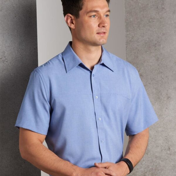 A man wearing a light blue short-sleeved button-up shirt stands against a gray textured wall. He has short dark hair and is looking to the side with his hands clasped in front of him.