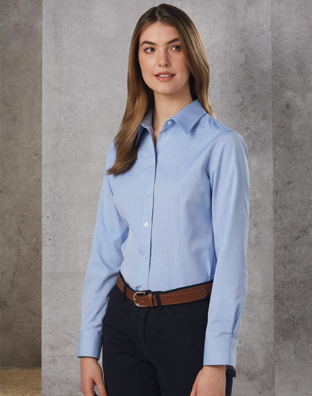 A person with long brown hair is wearing a light blue button-up shirt and dark pants, standing in front of a gray textured wall. They have a neutral expression and their hands are relaxed by their sides.