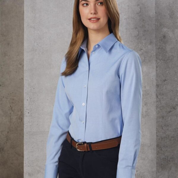 A person with long brown hair wearing a light blue button-up shirt, dark pants, and a brown belt stands against a textured gray wall. They are looking slightly to the side, with a neutral expression.