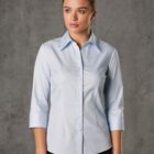 A woman in a light blue pinstripe shirt with three-quarter sleeves stands against a gray background. Her hair is neatly tied back, and she is looking slightly to the side.