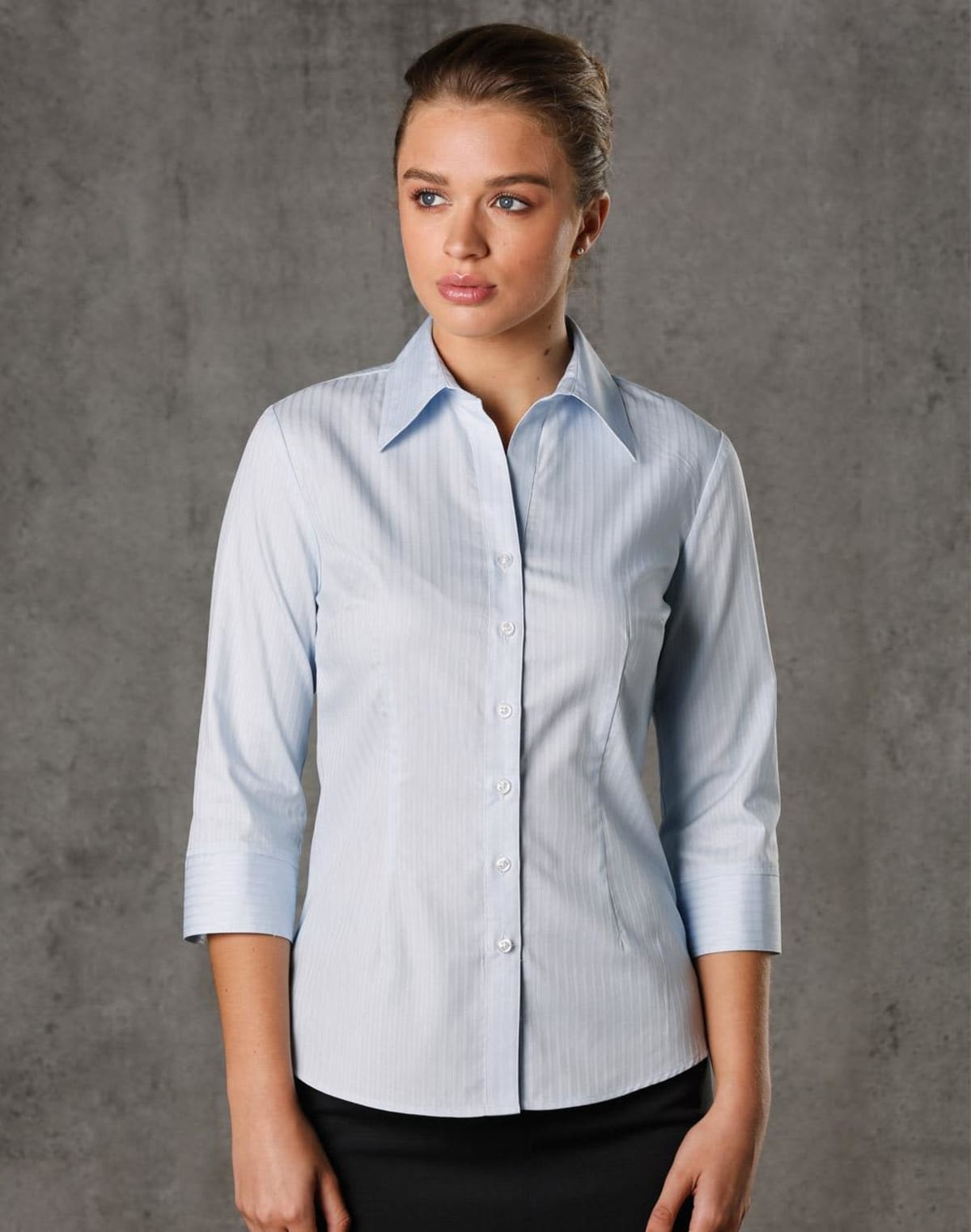 A woman in a light blue pinstripe shirt with three-quarter sleeves stands against a gray background. Her hair is neatly tied back, and she is looking slightly to the side.
