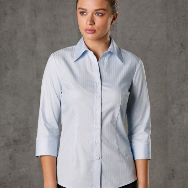 A person wearing a light blue button-up shirt with three-quarter sleeves stands against a gray background. They have short hair and a focused expression, looking slightly to the right.