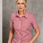 A person with blonde hair tied back is wearing a red and white checkered shirt with short sleeves. They are seated against a gray, textured background, looking directly at the camera.