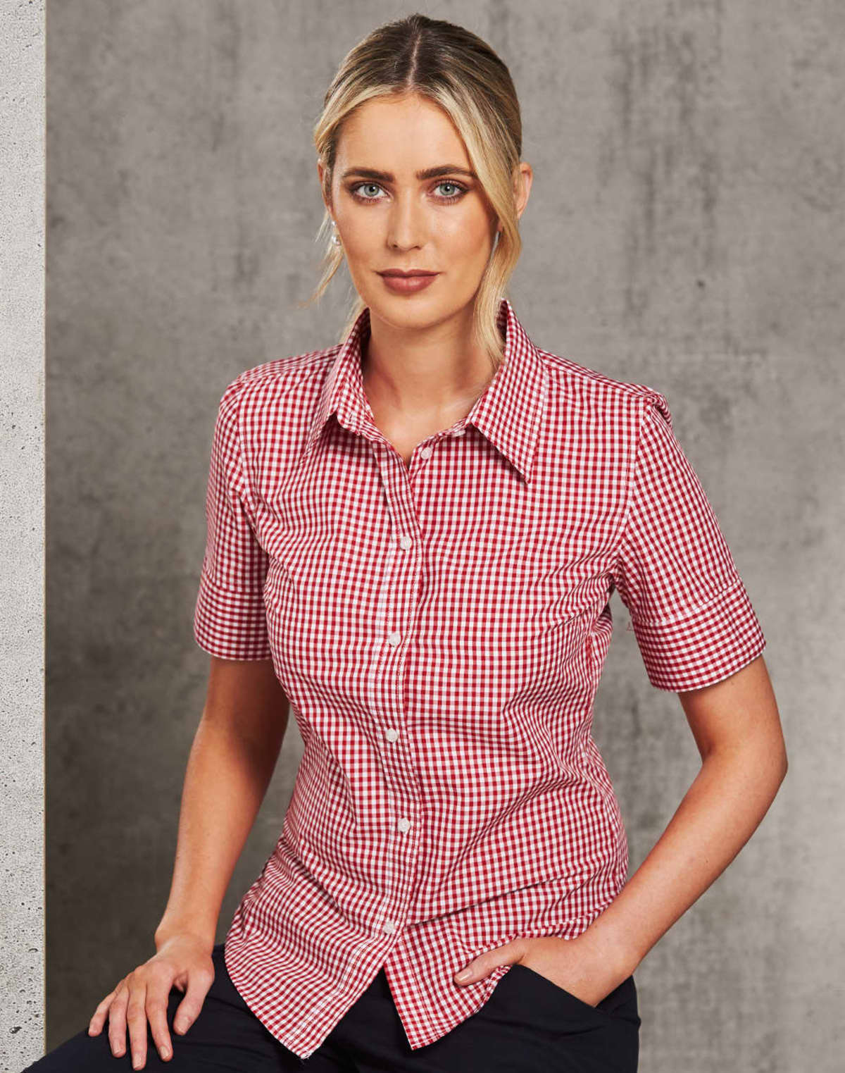 A person with blonde hair tied back is wearing a red and white checkered shirt with short sleeves. They are seated against a gray, textured background, looking directly at the camera.