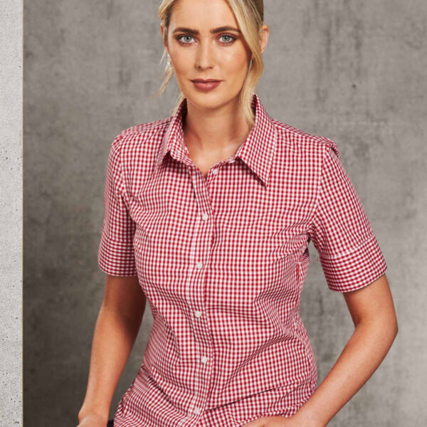 A person with blonde hair is wearing a red and white checkered shirt and dark pants, sitting against a gray concrete wall. Their expression is neutral.
