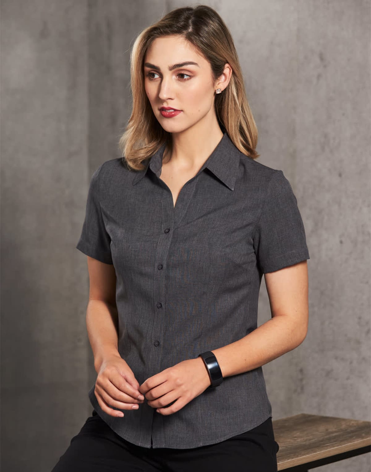 A woman with shoulder-length hair wears a dark gray, short-sleeve button-up shirt. She is standing in front of a gray concrete-like wall, with her hands gently clasped in front of her.