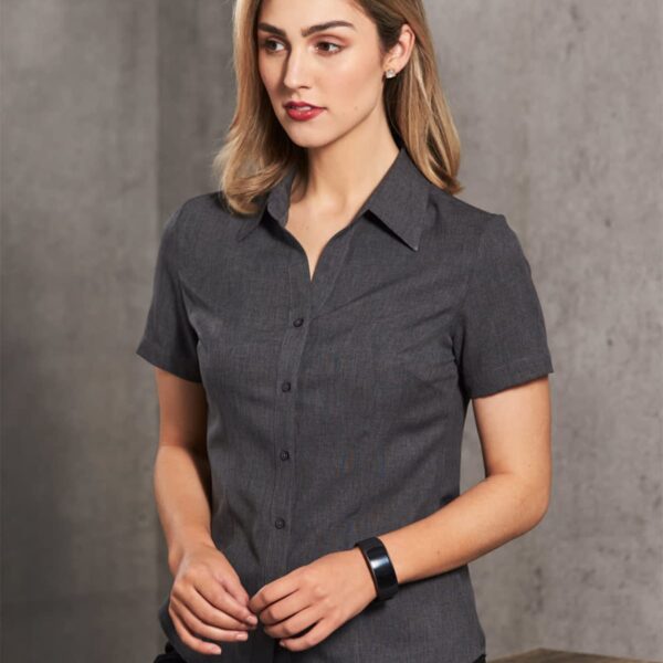 A woman with shoulder-length hair wears a gray short-sleeve button-up shirt, standing against a plain gray background. She looks to the side with a neutral expression and holds her hands together in front of her.