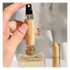 A hand holding a small, stylish perfume spray bottle with a wooden design. A larger bottle is in the background, and the hand has a pearl-accented nail. The setting is well-lit, highlighting the natural materials.