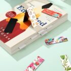 A book titled Educated by Tara Westover lies on a light green surface. Several colorful magnetic bookmarks, including floral, Christmas, and carnival themes, are clipped on and around the book.
