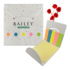 A set of colorful sticky notes in a recycled paper case with the text Bailey Nurseries. The case has five small colorful circles and is open to display sticky notes. Red flowers are in the background.