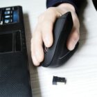 A hand using a vertical ergonomic mouse next to a laptop on a white desk. A small USB receiver lies beside the laptop. The setup is designed for comfortable computer use.