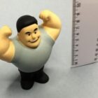 A small figurine of a smiling man flexing his muscles stands next to a ruler. The ruler measures about 8 centimeters, providing a scale for the figurines height. The figurine wears a gray shirt and black pants.