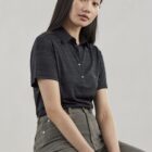 A person with long dark hair sits on a wooden stool against a neutral background. They are wearing a dark, short-sleeved polo shirt and gray pants, looking towards the camera with a calm expression.