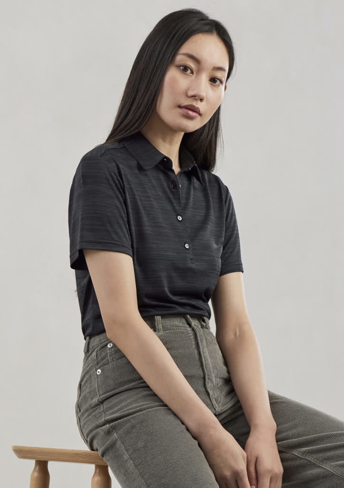 A person with long dark hair sits on a wooden stool against a neutral background. They are wearing a dark, short-sleeved polo shirt and gray pants, looking towards the camera with a calm expression.