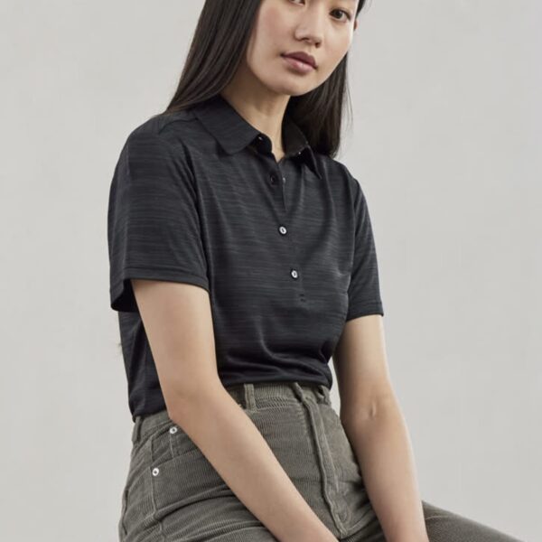 A person with long, dark hair sits on a wooden stool against a neutral background. They are wearing a black polo shirt and gray pants, looking directly at the camera with a calm expression.