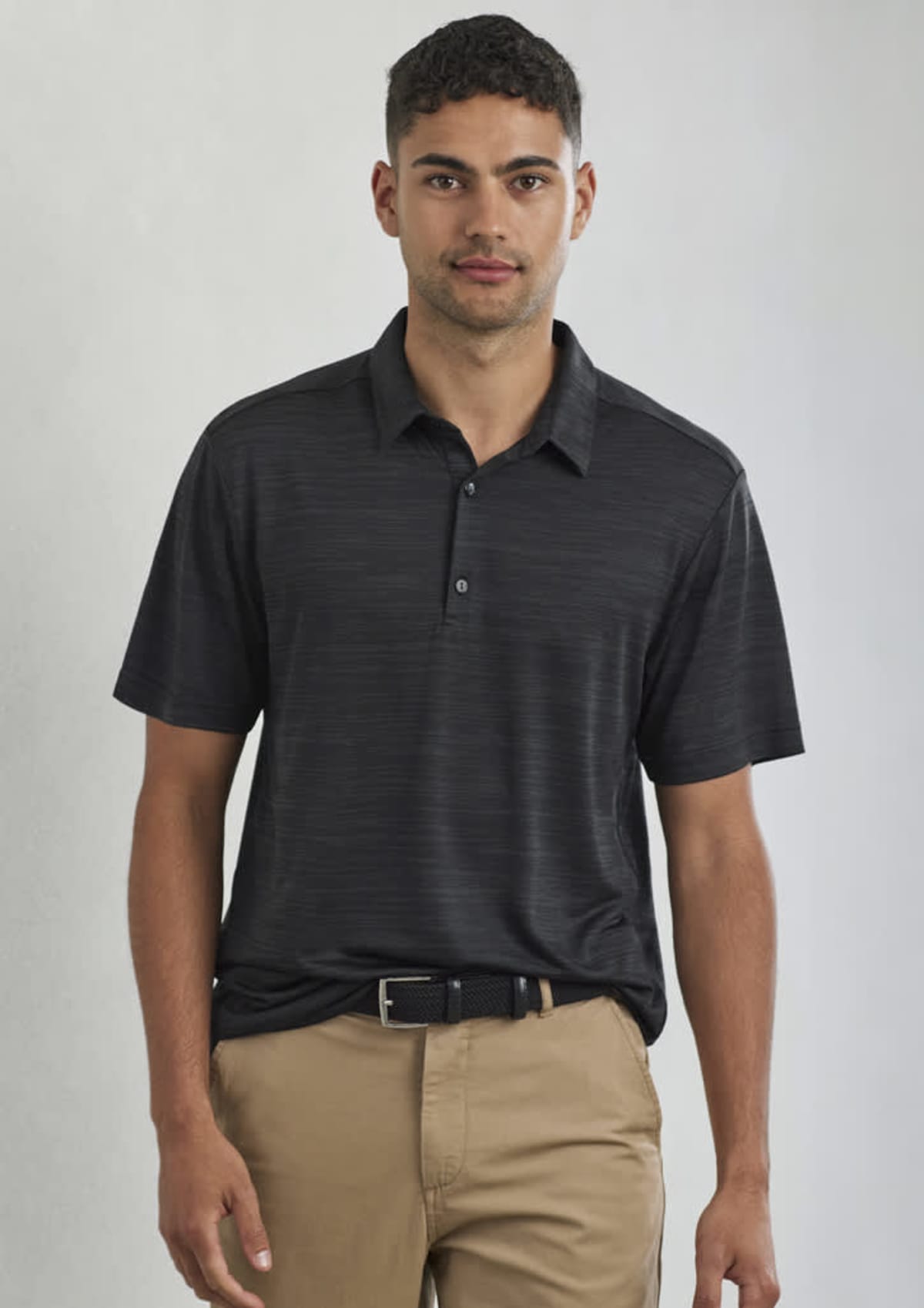 A man wearing a black polo shirt and tan pants stands against a plain background. He has short, curly hair and a neutral expression.
