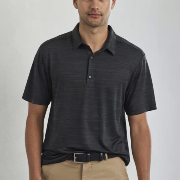 A man wearing a dark short-sleeve polo shirt and beige pants stands against a neutral background. He has short hair and a slight smile.