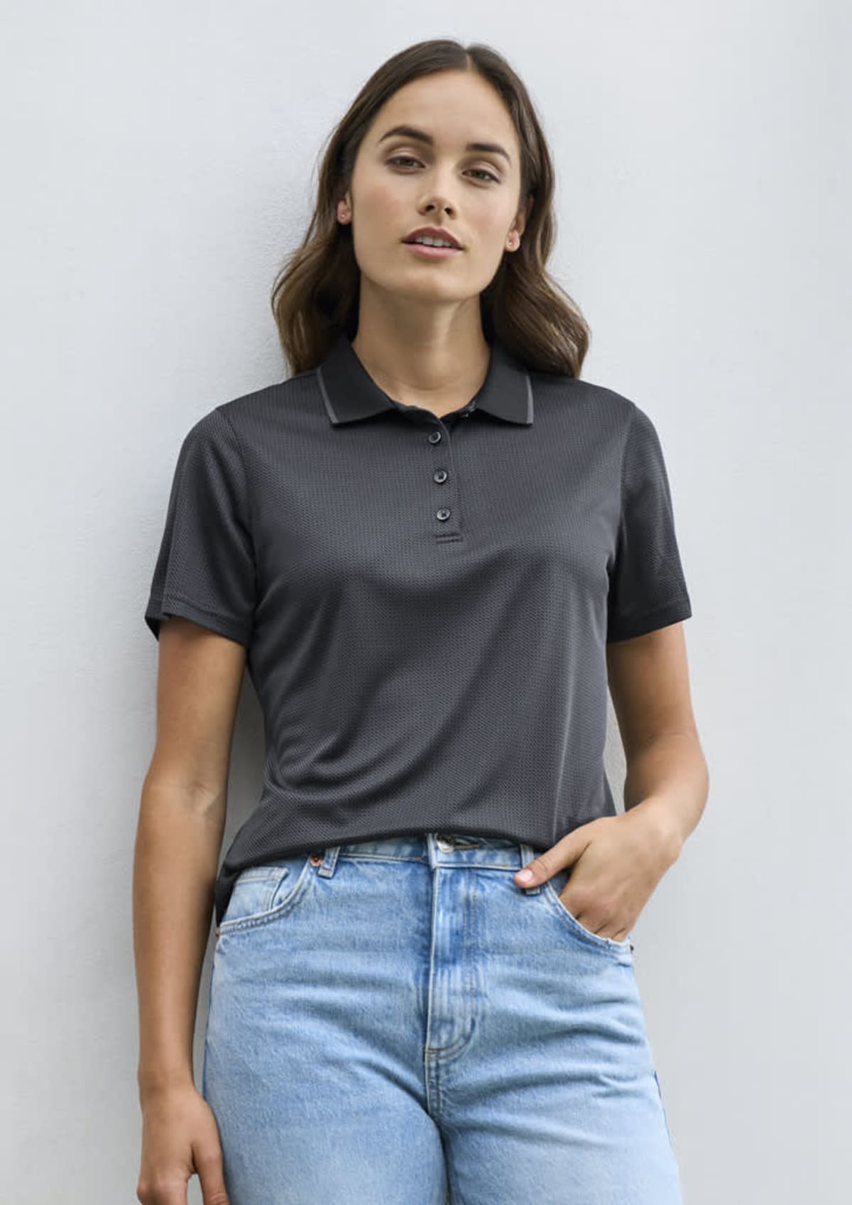 A person with long, dark hair wears a short-sleeved, dark polo shirt and light blue jeans, standing against a plain, light-colored wall.