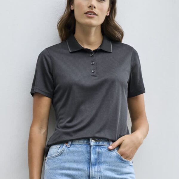 A woman with long brown hair wears a dark gray polo shirt and light blue jeans. She is standing against a plain white background, looking toward the camera with a relaxed expression.