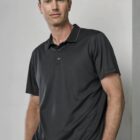 A man wearing a dark polo shirt stands against a light gray wall with hands in pockets, looking directly at the camera.