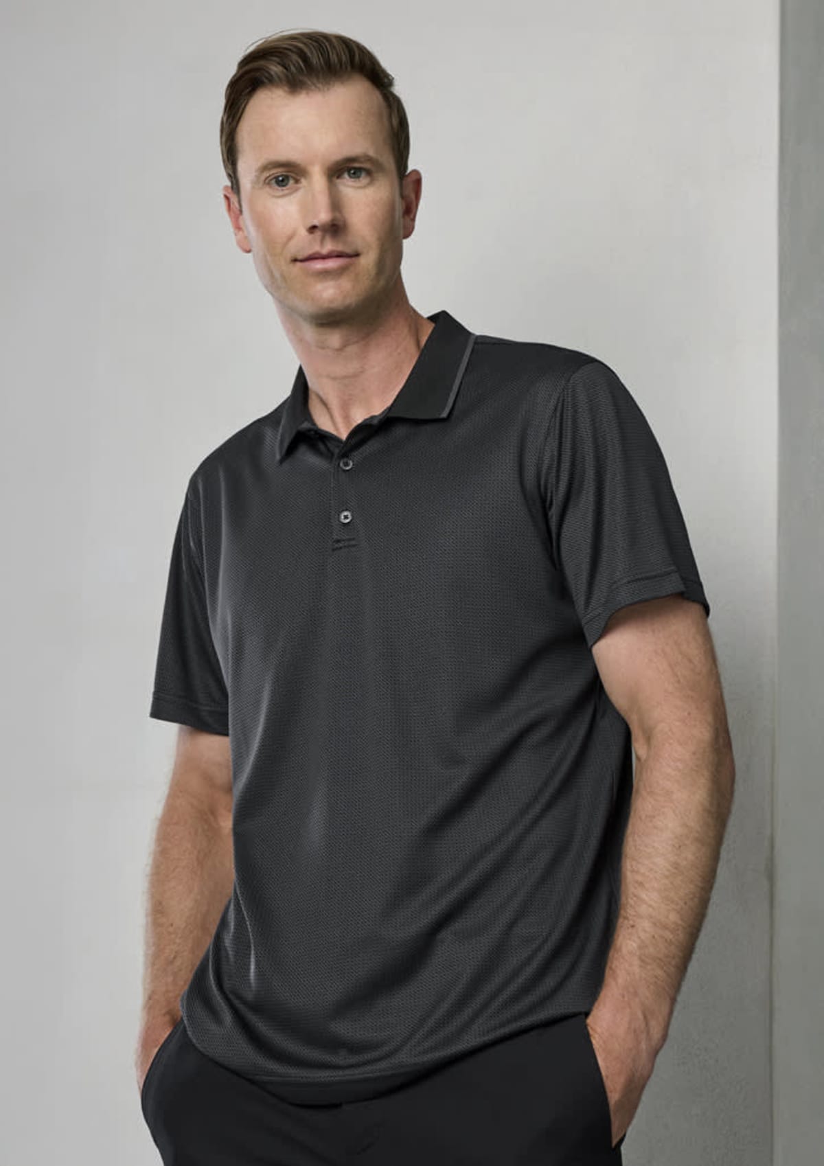 A man wearing a dark polo shirt stands against a light gray wall with hands in pockets, looking directly at the camera.