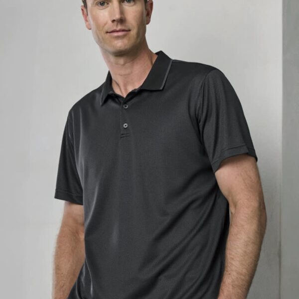 A man with short brown hair is leaning against a wall, wearing a dark polo shirt and black pants. The background is a neutral, light-colored wall.