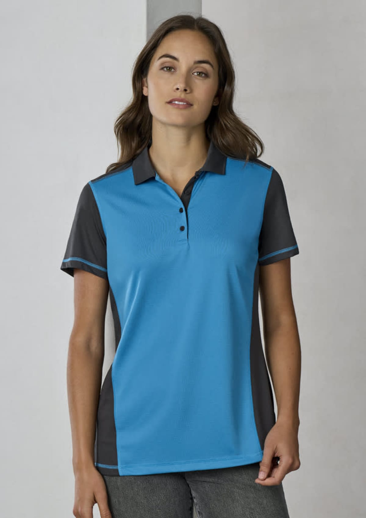 A person wearing a blue and dark gray polo shirt stands against a neutral background. They have long, dark hair and are looking directly at the camera.