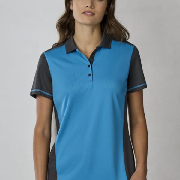 A woman with long hair wearing a blue and gray short-sleeve polo shirt and jeans stands against a plain background.