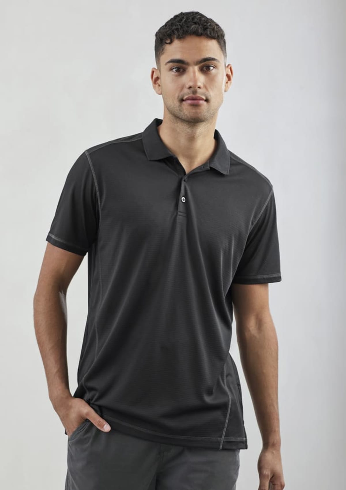 A man wearing a black polo shirt stands against a plain background. He has short curly hair and is looking at the camera with a neutral expression. One hand is in his pocket.