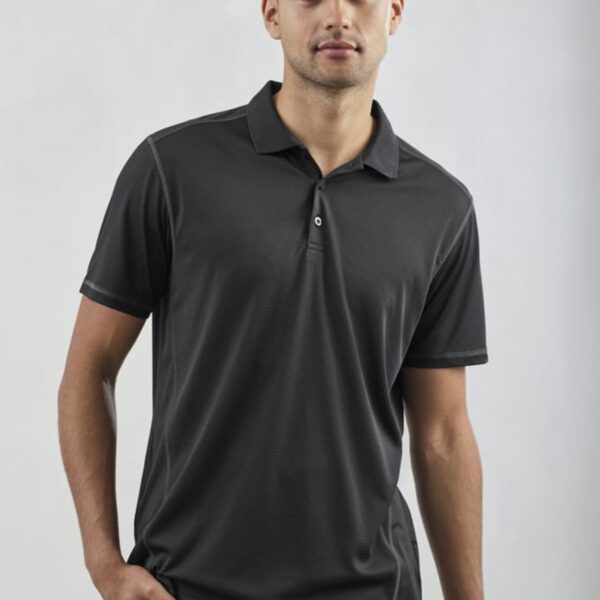 A person wearing a dark polo shirt and pants stands against a plain background, with one hand in their pocket.