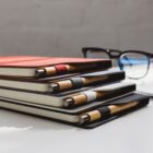 Stack of four notebooks with pens tucked in the spines, arranged on a white surface. Notebooks have black, red, and beige covers. A pair of glasses is placed in the background.