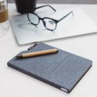 A gray notebook with a pen rests on a white desk beside a pair of glasses and a closed laptop. A small black plant pot slightly appears in the background.