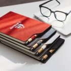A stack of four notebooks with pens in matching elastic holders. The top notebook is red with a logo, while the others are dark gray, gray, and white. A pair of black glasses and a laptop are in the background.