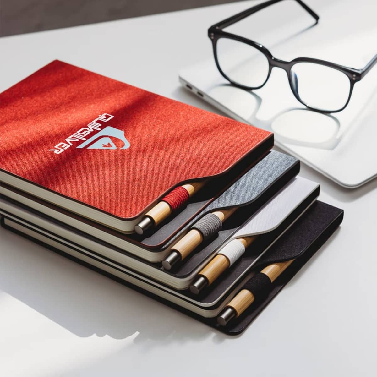 A stack of four notebooks with pens in matching elastic holders. The top notebook is red with a logo, while the others are dark gray, gray, and white. A pair of black glasses and a laptop are in the background.