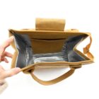 A hand holding open a tan insulated bag, revealing its silver-lined interior. The bag has handles and a small pocket inside. The exterior appears to be made of a textured material.