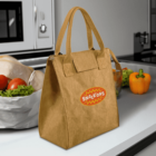 Brown tote bag with a Snackons logo sits on a kitchen counter. Behind it are vegetables, including lettuce, oranges, tomatoes, and a yellow pepper, next to a silver bowl containing a chicken. The room background is a modern kitchen setup.