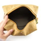 A persons hand holding an open, empty brown pouch with a zipper against a white background.