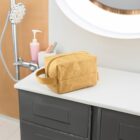A tan toiletry bag sits on a white bathroom countertop next to a gray cabinet. In the background, theres a pink liquid soap dispenser and a modern round mirror with a light.