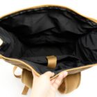 A hand holds open a brown backpack, revealing its spacious and empty black interior.