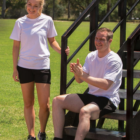 Two people in Premium Promotional T Shirts and black shorts enjoy a sunny day outdoors. One sits on grass-covered steps, clapping, while the other stands nearby. Their stylish attire complements the vibrant setting, highlighting a moment of casual elegance.