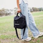 Person walking on grass, holding a black Patagonia lunch bag. Theyre wearing light blue jeans and a white shirt. Trees and sky are visible in the background.