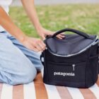 A person sitting on a striped blanket outdoors is zipping a black Patagonia insulated cooler bag. The person is wearing light blue jeans and a white shirt. Grass is visible in the background.