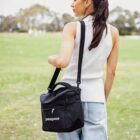 A woman stands outdoors on a grassy field, wearing a white sleeveless top and jeans. She carries a black Patagonia shoulder bag. Trees are visible in the blurred background.