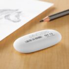 An oval white eraser labeled FACTIS OV 12 with a barcode, placed on a wooden surface next to a sharpened pencil and a sheet of paper featuring a pencil sketch.