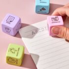 A hand places a colorful die labeled SANDWICH on a notepad with ABCDE written on it. Nearby are dice labeled PAPER, BASEBALL, and DUCKY STEPS. The background is a soft pink surface.
