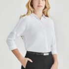 A person with long blonde hair is wearing a white button-up shirt and black pants, standing with one hand in their pocket. The background is plain and light-colored.