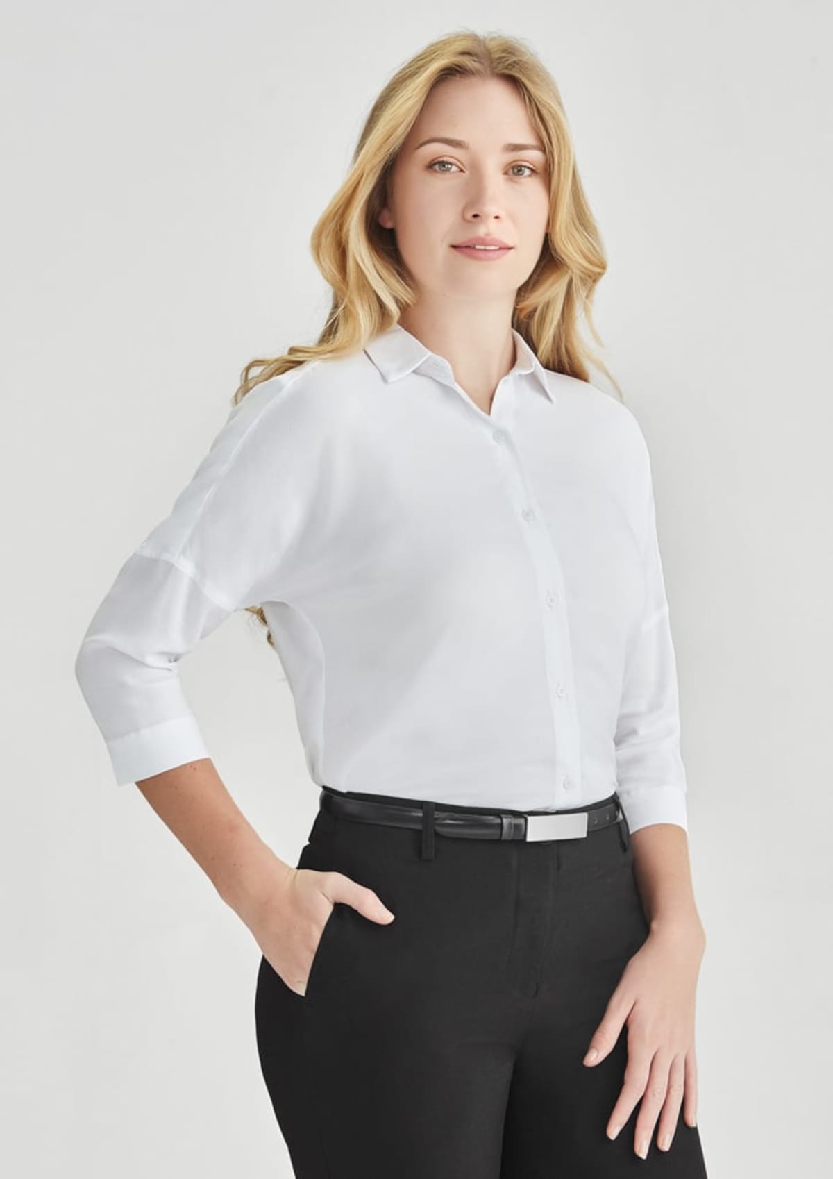 A person with long blonde hair is wearing a white button-up shirt and black pants, standing with one hand in their pocket. The background is plain and light-colored.