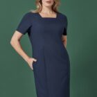 A person stands confidently wearing a navy blue, short-sleeved dress with a square neckline. Their hand is in a pocket, and they pose against a solid green background.